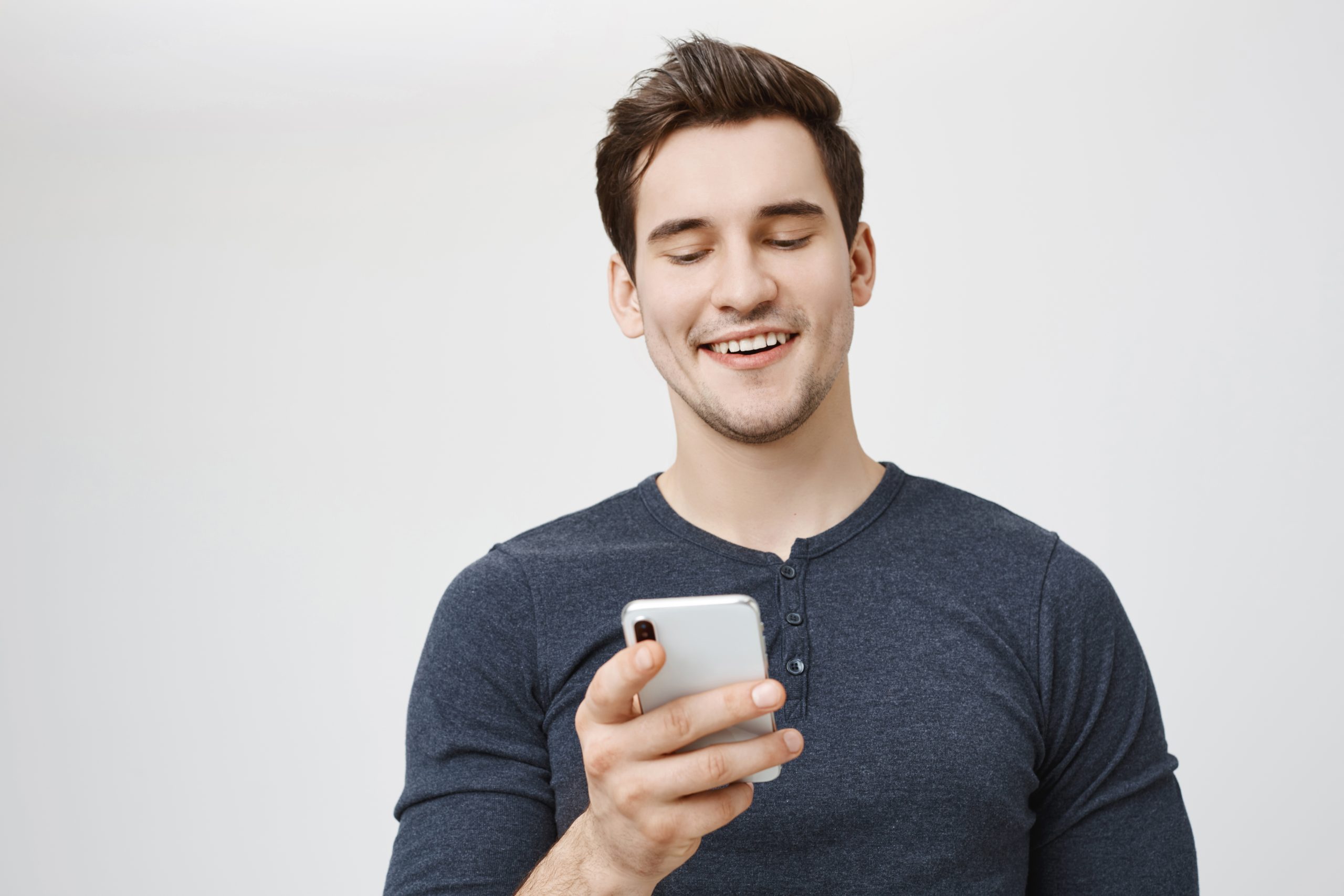 European dark-haired man in casual clothes posing against gray background having happy expression while reading messages from friends, smiling joyfully. Modern communication and technologies
